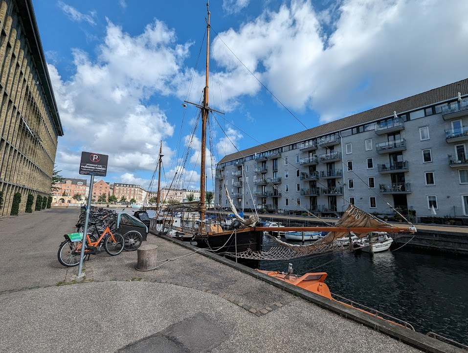 A scenic waterfront view featuring a tall sailing ship docked in a canal. In the foreground, there is a bike rack with several bicycles, including an orange bike. On the left, a sign indicates parking restrictions, and on the right, modern apartment buildings with balconies overlook the water. The sky is partly cloudy, creating a bright and inviting atmosphere.