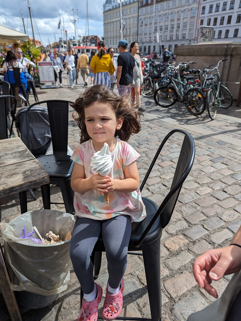 A young girl with curly hair sits on a black chair, holding a large soft-serve ice cream cone. She wears a tie-dye shirt, gray leggings, and pink crocs. In the background, there are people walking along a cobblestone path, with bicycles parked nearby and buildings lining the street under a partly cloudy sky. A trash can is visible next to her.