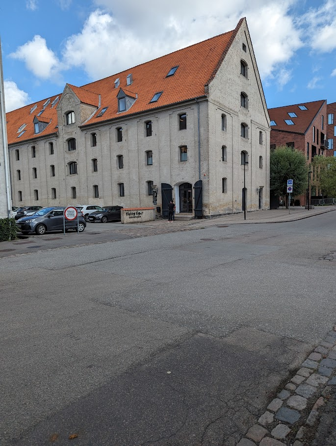 A large, rectangular building with a light-coloured façade and a distinctive red-tiled roof is seen on the corner of a street. The building features multiple windows and a main entrance with a sign that reads "Flying Tiger, Copenhagen". Several parked cars are visible in front of the building, and the sky above is partly cloudy. A sidewalk and street signage are also present in the foreground.