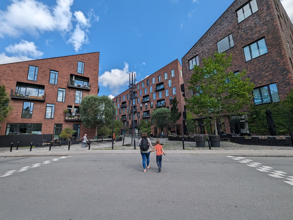A woman and a child walk hand in hand on a paved street surrounded by modern brick apartment buildings. The sky is blue with scattered clouds. The area features greenery and some outdoor seating areas. In the background, a few people are seen walking in different directions.