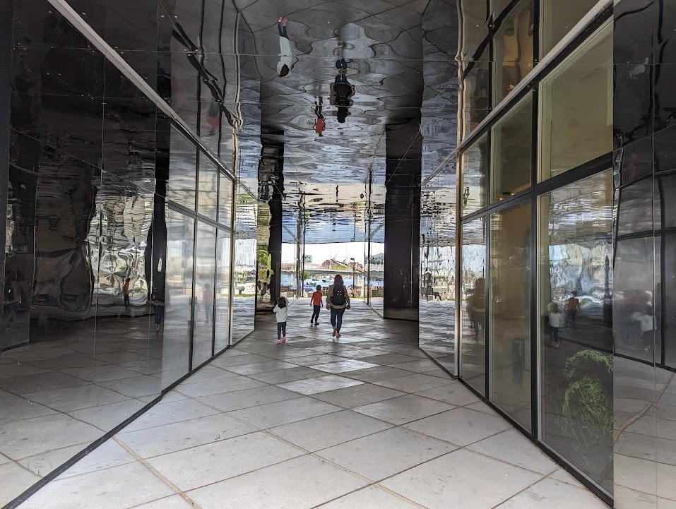 A group of children and an adult walk through a modern, reflective corridor with sleek black walls and large glass windows. The ceiling is also reflective, creating a distorted view of the space above. In the background, there is natural light coming from outside, illuminating the area.