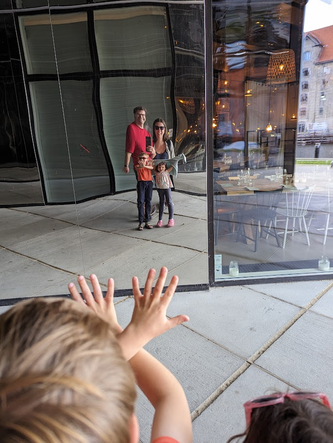 A family of four stands in front of a reflective building facade, smiling for a selfie. The parents, a man in a red shirt and a woman in a gray jacket, hold their two children, a boy and a girl. In the foreground, two children with their backs to the camera extend their hands towards the family. The scene is set in an urban environment with modern architecture and warm lighting visible through the glass.