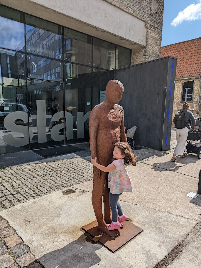 A young girl with curly hair is playfully hugging a life-sized sculpture of a standing figure made of rusted metal. The sculpture is situated outside a modern building with large glass windows. The scene is set on a sunny day, with people walking in the background. The girl wears a colourful shirt and pink shoes.