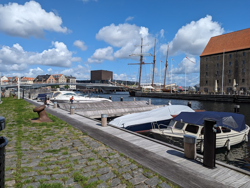 A scenic view of a marina with several boats moored along a wooden dock. In the background, there are modern buildings and a tall ship with masts. The sky is clear with fluffy white clouds, and the area features grassy patches and cobblestone paths along the water.