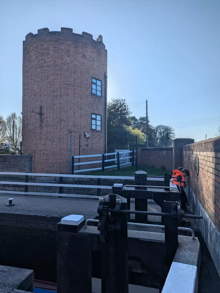 A brick tower with a rounded top (the Gailey Roundhouse) stands beside a canal. In the foreground, a lock mechanism is visible, and two children in orange life jackets are preparing to close one side of the lock gates. The lock gate is designed at a right angle because it is so close to the road bridge. The scene is bright under clear blue skies.