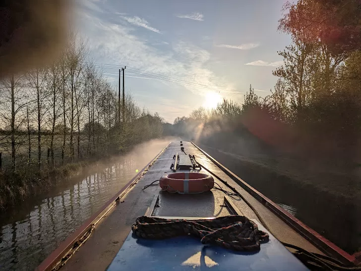 A canal boat is traveling along a misty waterway, surrounded by trees and power lines. The sun is rising in the background, casting soft light over the scene, creating a serene and tranquil atmosphere. A life buoy and ropes are visible on the boat's deck.