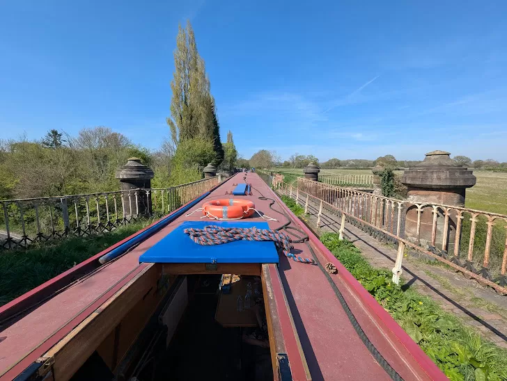A view from the top of a canal boat, showcasing a blue cover, orange life buoy, and a length of chain. In the background, tall trees line a path, and fence borders the canal as it crosses over an aqueduct, against a clear blue sky. The setting appears peaceful and rural.