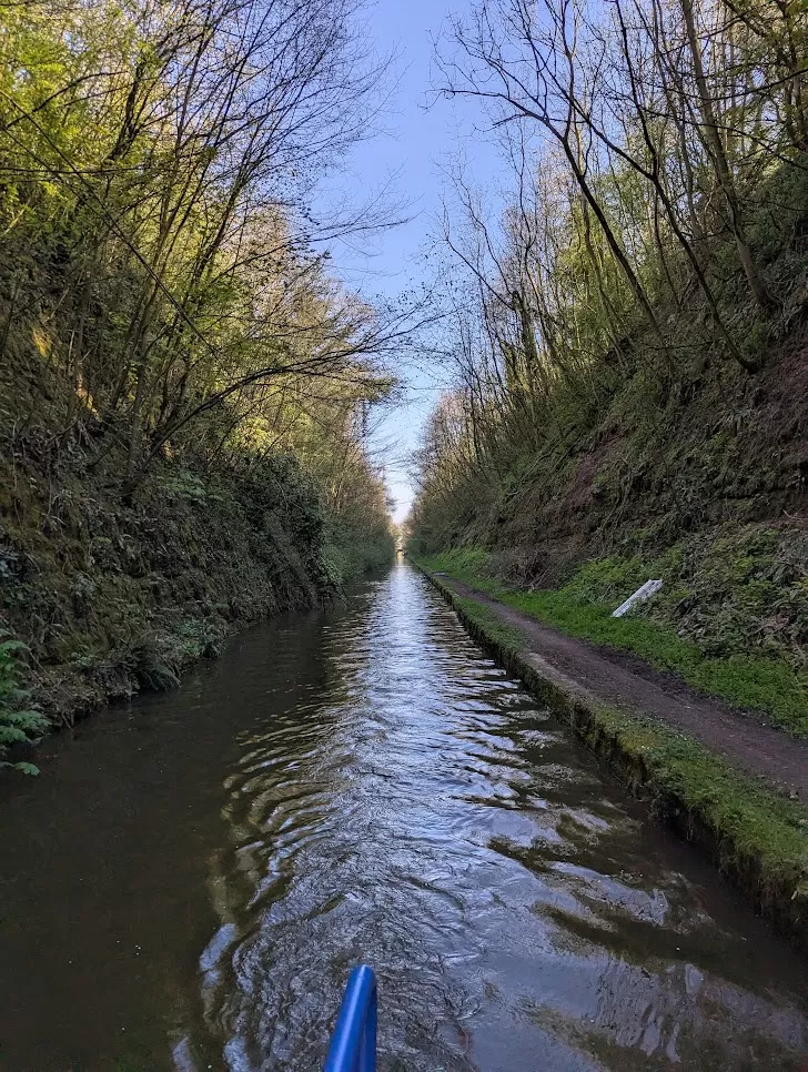 A narrow, tranquil canal surrounded by lush green trees and steep, natural banks. The water is gently rippling under a clear blue sky, with a path running alongside the right side of the canal. The perspective looks down the length of the canal, creating a sense of depth.