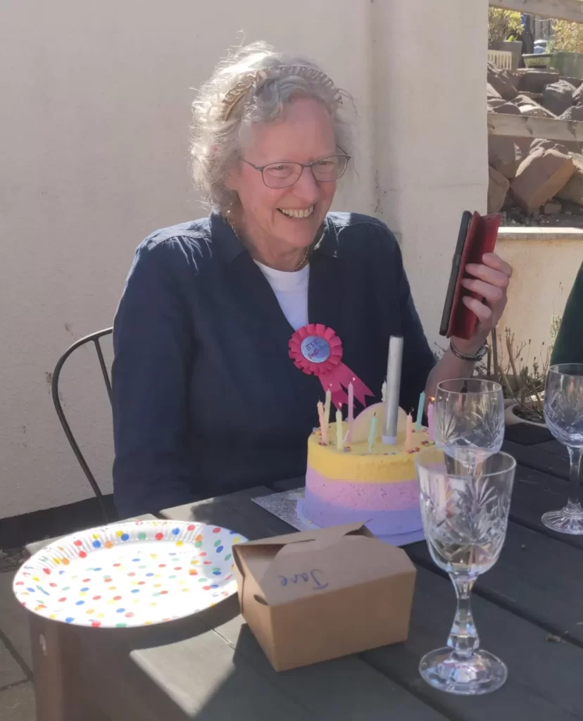 A smiling older woman with curly gray hair is sitting at a table outdoors, celebrating a birthday. She is wearing a blue shirt and a pink birthday ribbon. In front of her is a colorful birthday cake with candles, a decorated plate, a box, and several glasses. She is holding a phone and appears joyful during the celebration.