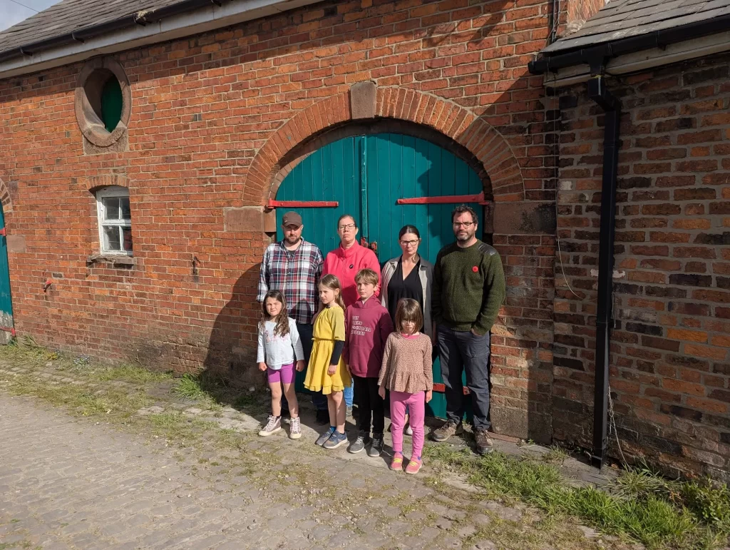 A group of eight adults and children stands in front of a rustic brick building with a large teal door. The adults are casually dressed, with one wearing a plaid shirt and another in a green sweater. The children are in colourful outfits, smiling at the camera. The scene is set outdoors on a sunny day, with green grass and a cobblestone path visible.
