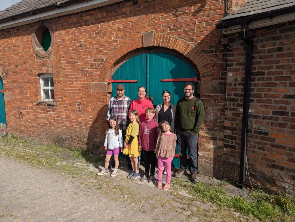 A group of seven people, including three children, stands in front of a rustic brick building with a large teal door. The group is smiling, dressed casually, and appears to be enjoying a sunny day outdoors.