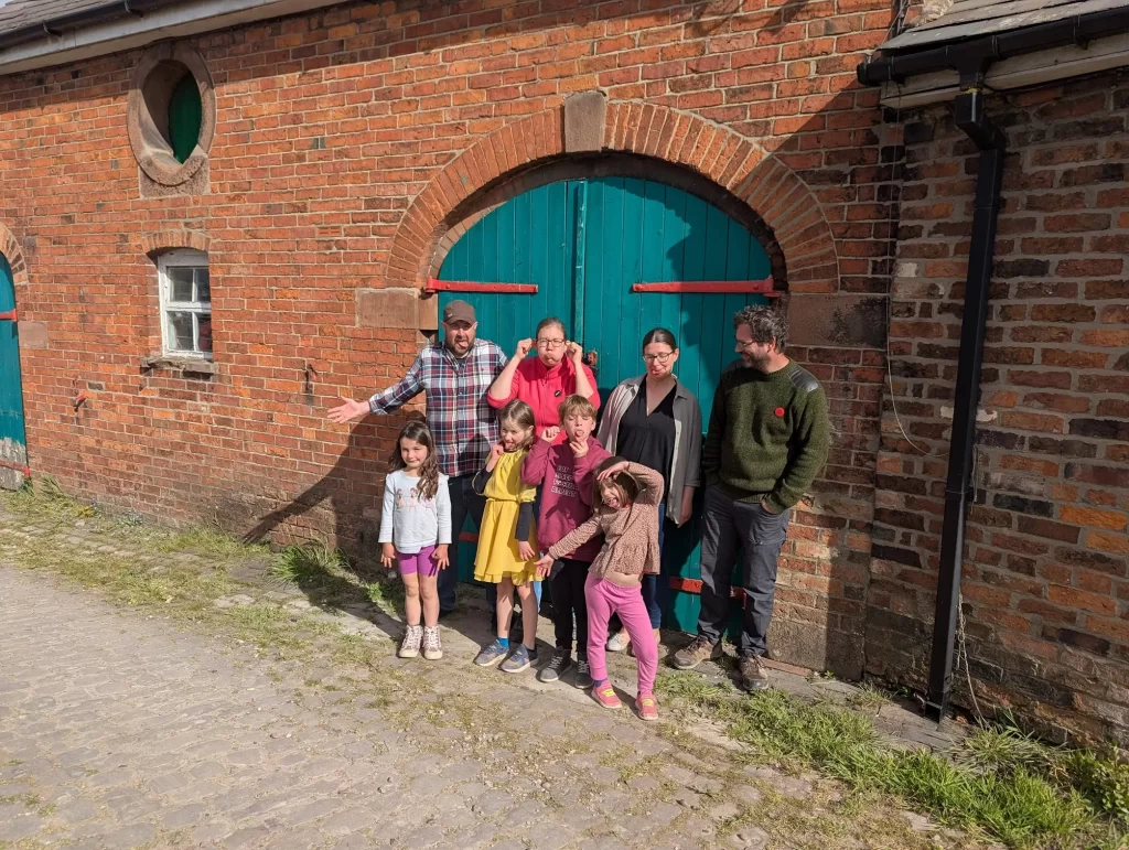 A group of eight people, including four children, poses in front of a rustic red-brick building with a large blue-green double door. The adults are dressed casually, while the children wear colourful summer outfits. They are smiling and showing playful poses, creating a cheerful family atmosphere.