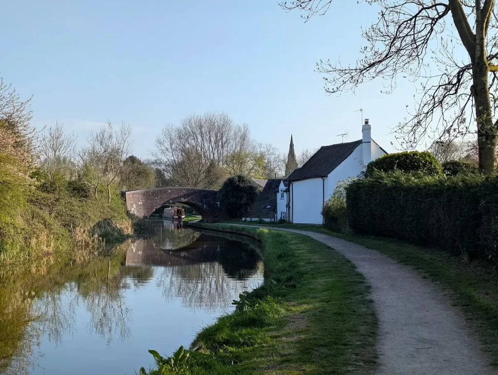 A tranquil scene featuring a winding path alongside a calm canal, bordered by lush greenery. In the background, a quaint white house and a stone bridge can be seen, with a church spire visible above the trees. The sky is clear and blue, suggesting a sunny day.