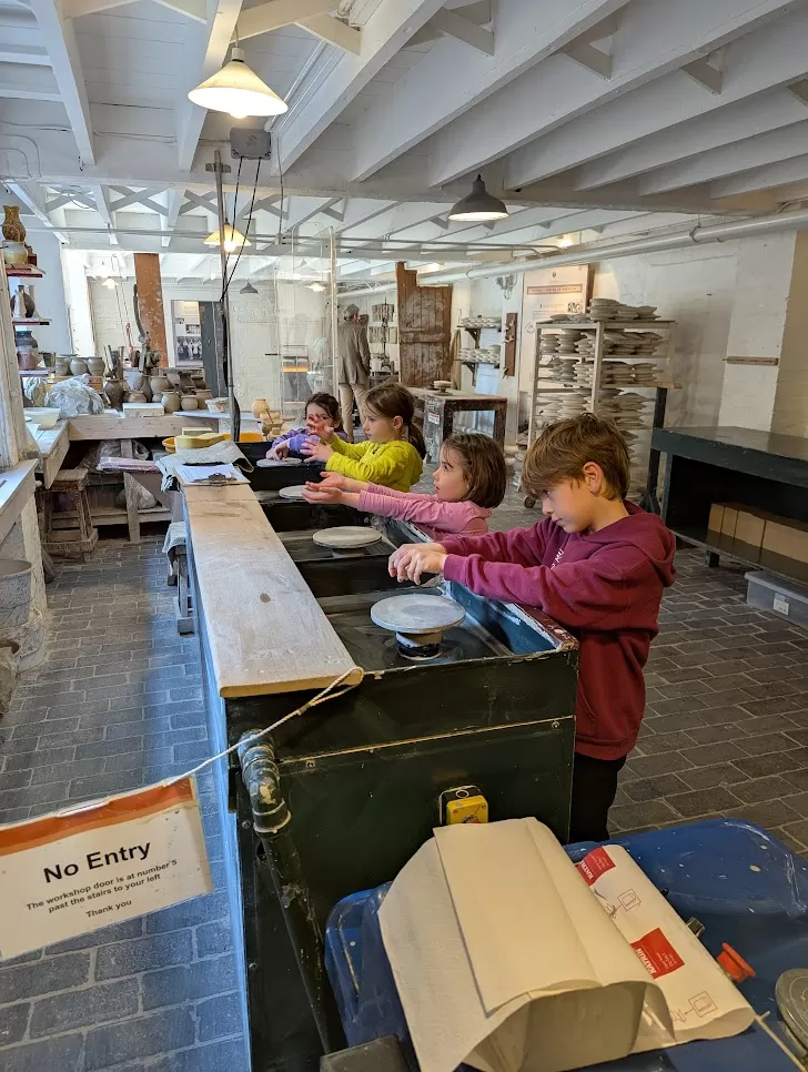 Four children participate in a pottery class, each working at a pottery wheel. They are concentrated and engaged, wearing colorful clothing in a bright, spacious studio. A sign reading "No Entry" is visible nearby, indicating the area might be restricted. Shelves filled with pottery supplies and finished pieces are in the background.