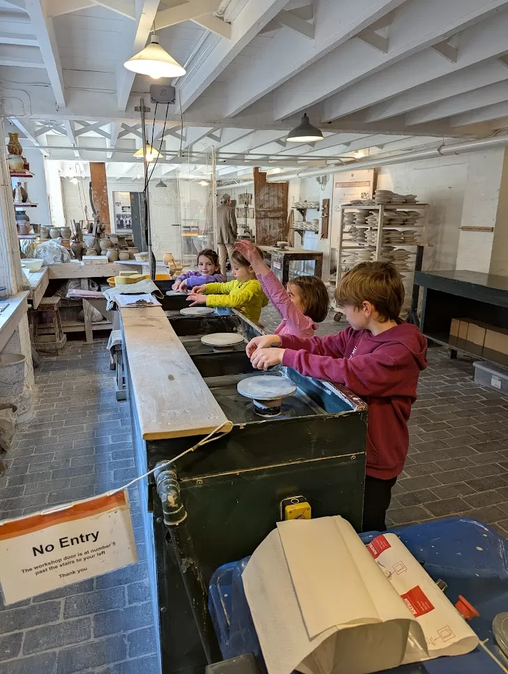 Four children are engaged in a hands-on activity at a worktable in a bright, spacious room. They are interacting with materials, with equipment and tools visible around them. A "No Entry" sign is prominently displayed in the foreground, indicating restricted access. The setting resembles a workshop or educational space designed for creative projects.