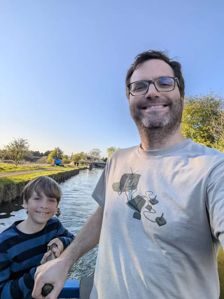 A man and a boy are smiling while taking a selfie on a boat in a calm waterway. The background features greenery and a clear blue sky. The boy is wearing a striped shirt, and the man is in a t-shirt with a graphic design.