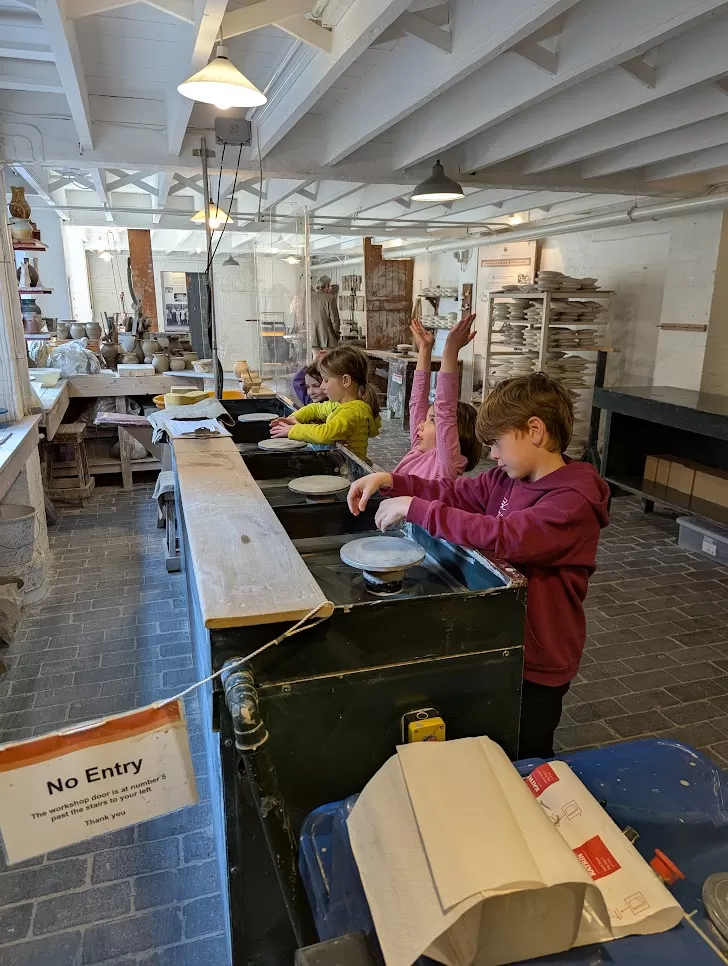 Four children are engaged in a hands-on activity inside a workshop. They are standing at a long workbench, each focused on a task, while one child raises their hand enthusiastically. The room is bright and filled with various tools and materials, with a "No Entry" sign visible in the foreground.