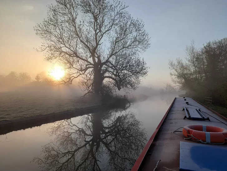 A misty morning scene with a calm canal reflecting the early sunlight. A solitary tree stands on the riverbank, casting a shadow on the water. A narrowboat is visible on the right side, with a bright orange life ring on its roof. The atmosphere is tranquil, with soft colours of dawn.